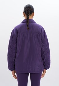Paarse gewatteerde jas met een ontspannen pasvorm, voorzien van een kielkraag en elastische manchetten. Het materiaal heeft een gladde, glanzende textuur.