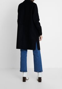 Černý vlněný kabát s jednoduchým designem a dlouhými rukávy, s bočními štěrbinami, v kombinaci s modrými rozšiřujícími se džínami a bílými botami na podpatku.