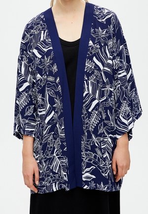 Person iført en løs, mørkeblå kimono med hvide tropiske blad- og blomsterprint over en sort top.