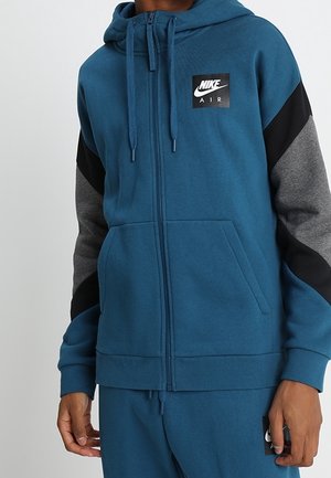 Mężczyzna noszący niebieską bluzę z kapturem Nike Air na zamek, z czarnymi i szarymi rękawami w blokach kolorów oraz pasujące spodnie.