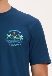 Tamsiai mėlynos spalvos medvilninė marškinėliai su spalvotu raštu, atvaizduojančiu palmės, bangas ir saulę, su užrašu "Antigua Port Beach Club" virš dizaino.