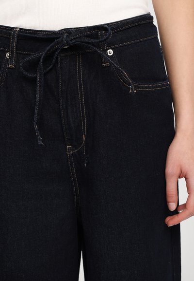 Mørke jeans med snøring i livet, synlig søm i kontrastfarge, og to frontlommer. Glatt tekstur og elastisk midje.