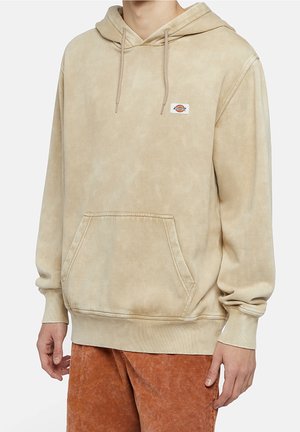Beige huppari, jossa etutasku ja pieni logopatch rintakehässä, käytetty ruosteenväristen housujen kanssa.