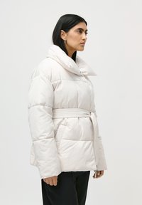 Femeie cu păr drept și negru poartă o geacă oversized albă, pufos cu guler înalt, curea și buzunare laterale, asortată cu pantaloni negri, întorsă spre dreapta.