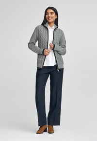 Grå kabelstickad cardigan med ståkrage och dragkedja, accentuerad med marinblå detaljer. Bärs över en vit skjorta och tillsammans med mörka byxor.