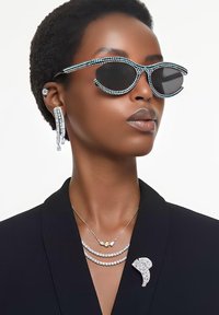 Schwarze Sonnenbrille mit Perlenakzenten, geschichtete silberne Halsketten, übergroße Ohrringe und eine dekorative Brosche an einem schwarzen Blazer.