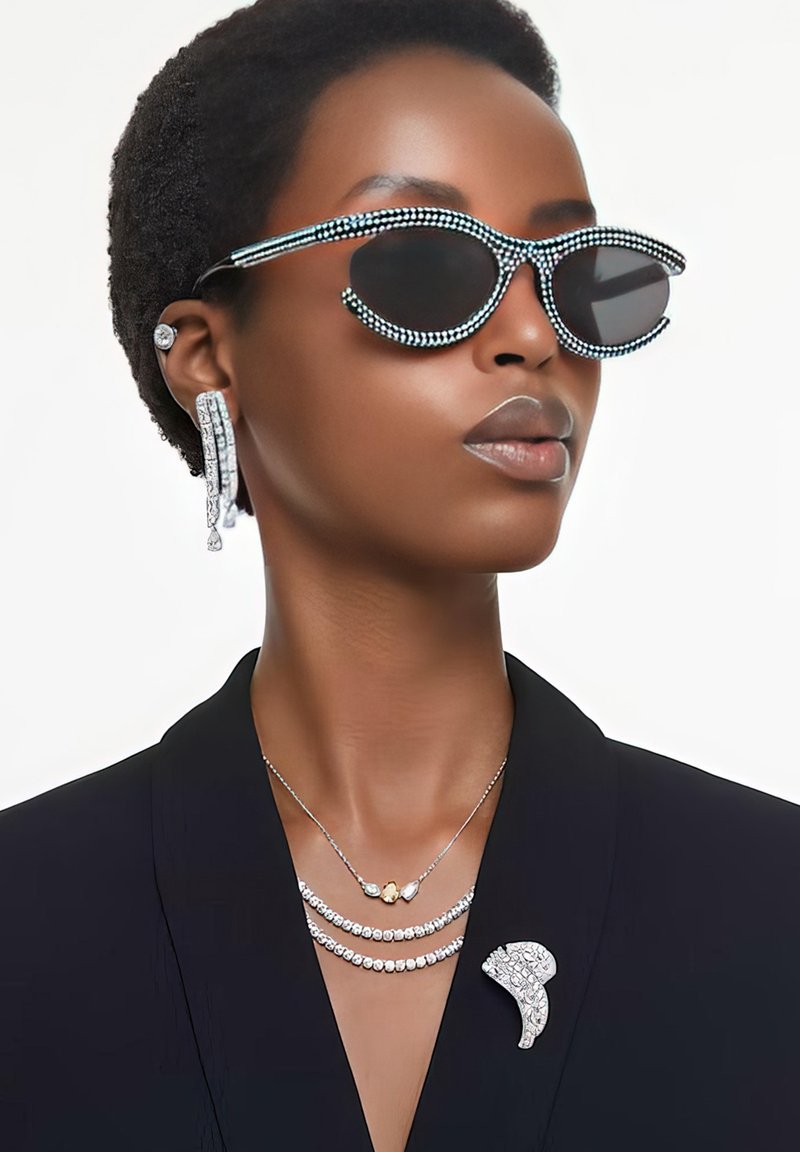 Schwarze Sonnenbrille mit Perlenakzenten, geschichtete silberne Halsketten, übergroße Ohrringe und eine dekorative Brosche an einem schwarzen Blazer.