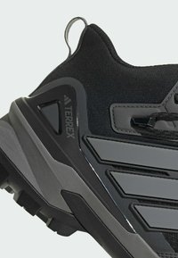 Chaussure de sport noire avec une tige en tissu texturé, un talon renforcé et des accents gris contrastants. Elle dispose d'une semelle en caoutchouc durable pour une meilleure adhérence.