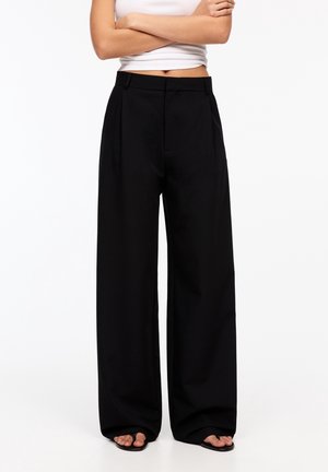 Persoon met zwarte wijde broekspijpen en zwarte sandalen met open tenen, armen gekruist, met een witte cropped mouwloze top tegen een effen achtergrond.