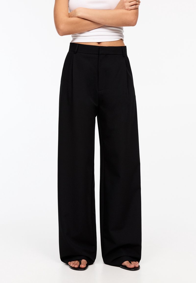 Personne portant un pantalon noir à jambes larges et des sandales noires à bouts ouverts, les bras croisés, avec un haut court blanc sans manches sur un fond uni.