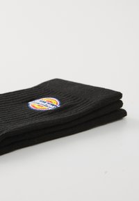 Sosete negre cu striații, aranjate ordonat, având un logo circular cu culorile roșu, galben și albastru pe o parte. Fundal simplu și minimalist.