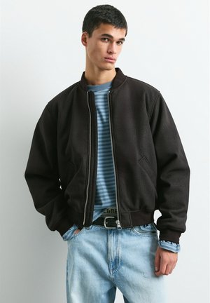 Un jeune homme porte une veste noire zippée par-dessus une chemise bleue à rayures et un jean bleu clair, sur un fond uni.