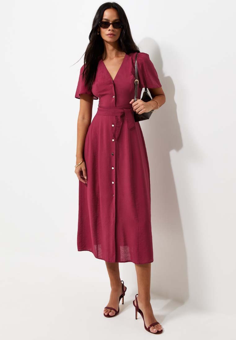 Woman wearing a buttoned, belted burgundy midi dress, dark sunglasses, heeled sandals, holding a small black handbag, standing against a white wall.