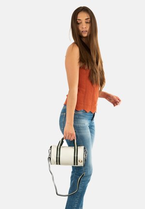 Borsa cilindrica bianca con manici neri. Texture liscia, con un piccolo emblem. Il modello indossa una maglietta arancione e jeans blu, tenendo la borsa.