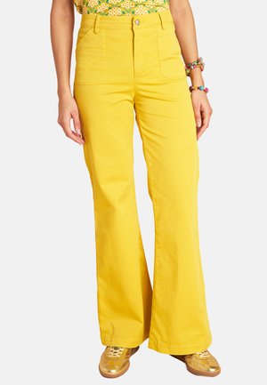 Frau trägt leuchtend gelbe, hoch taillierte und weit geschnittene Hosen, goldene Sneakers, geblümtes Oberteil und bunte Perlenarmbänder vor weißem Hintergrund.