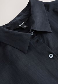 Chemise en lin bleu marine foncé avec col pointu, boutons blancs et étiquette de marque visible "NEXT" à l'intérieur du col.