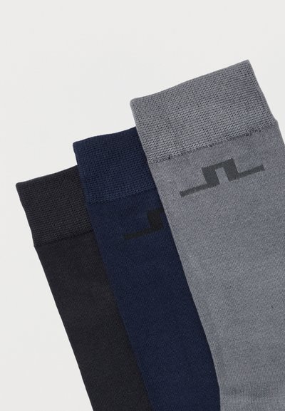 Τρία διπλωμένα ζευγάρια κάλτσες σε μαύρο, σκούρο μπλε και γκρι, το καθένα με υφασμένο γεωμετρικό λογότυπο κοντά στη μανσέτα.