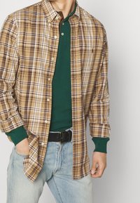 Camicia a quadri marrone e crema con bottoni sopra una maglietta verde a maniche lunghe; jeans di denim; cintura nera; presenta un design con bottoni e colletto pieghevole.