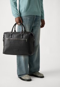Svart skinnveske med teksturert mønster, doble håndtak, glidelås og sølvfargede detaljer; båret av en person i denim.