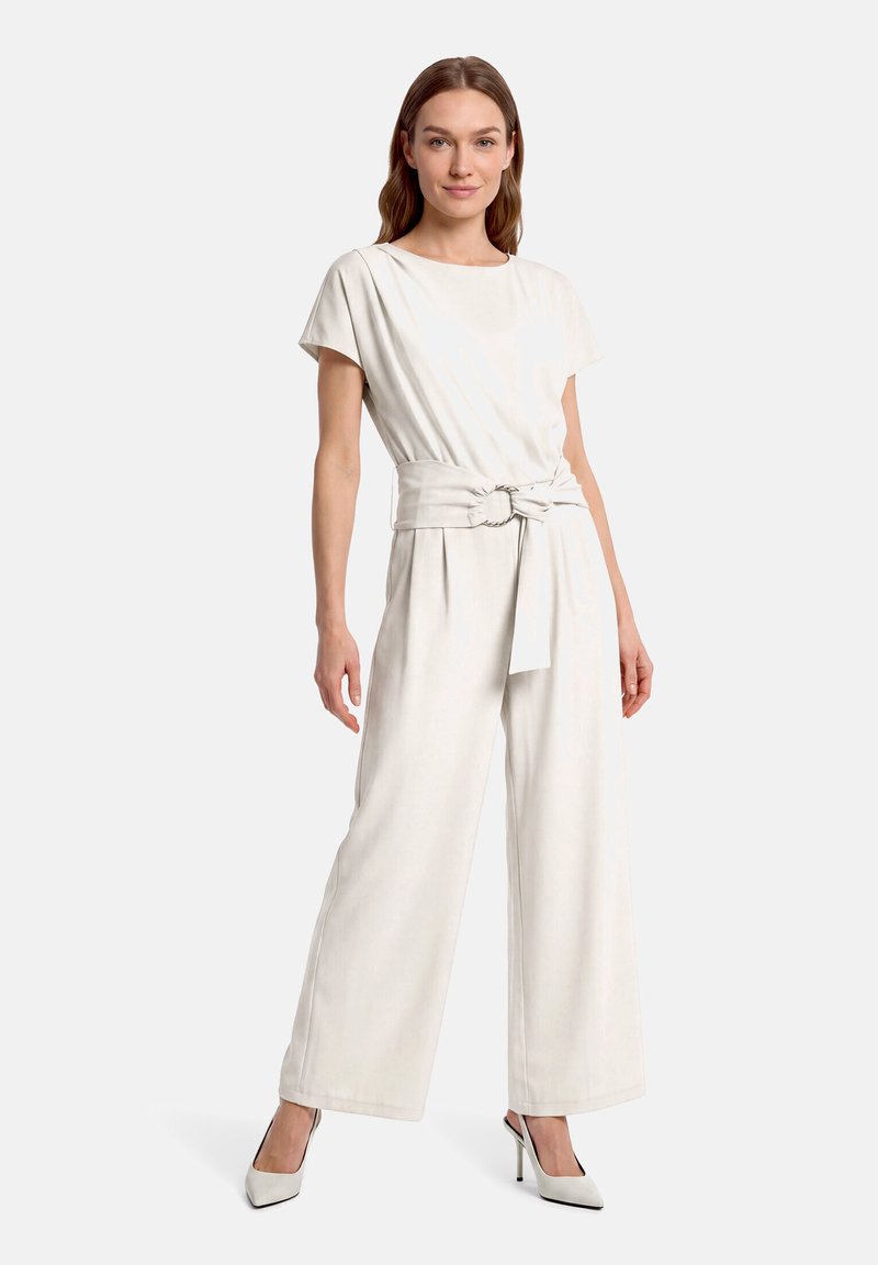 Femme debout portant une combinaison blanche à manches courtes avec une ceinture large et une boucle argentée, assortie à des chaussures blanches à talons hauts.