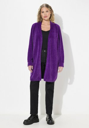 Cardigan violet peluche avec un devant ouvert, des manches longues et un ourlet côtelé. Porté par-dessus un top noir et associé à un pantalon noir et des bottes noires à semelles épaisses.
