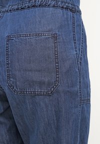 Gros plan d'un pantalon en jean bleu montrant une poche arrière cousue et des détails de couture sur le côté.