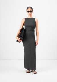 Kvinne i ermeløs, teksturert mørkegrå maxikjole som holder en stor svart kuvertveske, iført svarte solbriller og flate sandaler med slangeskinnmønster, hvit bakgrunn.