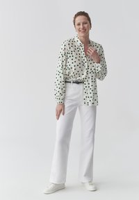 Camicia bianca con pois verdi, design button-down, maniche lunghe; indossata con jeans bianchi a zampa d'elefante e sneakers bianche. Dettaglio cintura semplice.