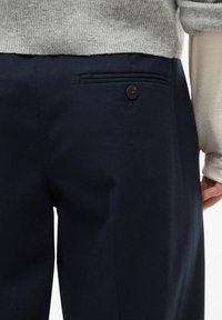 Pantalon bleu marine à texture lisse, avec une poche arrière munie d'un bouton noir. Le vêtement est partiellement couvert par un haut gris clair.