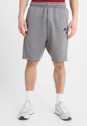 Mężczyzna noszący szare spodenki Nike, białe skarpetki oraz białe buty sportowe stoi z rękami swobodnie opuszczonymi wzdłuż ciała.