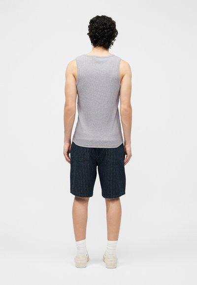 Άντρας με σγουρά μαύρα μαλλιά, ντυμένος με γκρι αμάνικο πουκάμισο, μπλε σκούρα σορτς με ρίγες, λευκές κάλτσες και μπεζ αθλητικά παπούτσια, στέκεται με την πλάτη προς τη μπροστινή πλευρά.