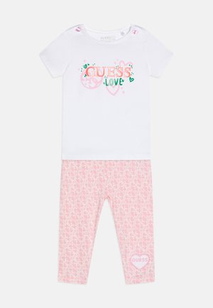 T-shirt bianco a maniche corte con grafica "GUESS LOVE", abbinato a leggins rosa con motivo a logo ripetuto e dettaglio a forma di cuore.