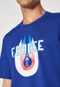 Zilas kokvilnas T-krekls ar uzrakstu "FRANCE" treknā baltā tekstā, akcentēts ar grafiku sarkanā un zilā krāsā, kā arī Adidas logo centrā.