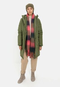 Olijfgroene pufferjas met een capuchon, roze en grijs geruite sjaal, beige trui, crèmekleurige broek en taupe veterschoenen.