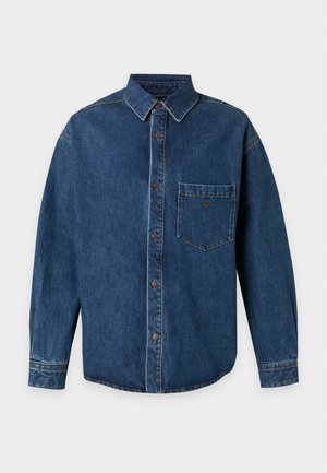 Mann trägt übergroßes blaues Jeanshemd mit Knopfleiste, dunkle Hose und schwarze Schuhe und steht vor einem schlichten hellen Hintergrund.