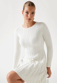 Robe blanche en tricot à manches longues, avec une texture côtelée et une taille cintrée, s'évasant légèrement au niveau de l'ourlet.