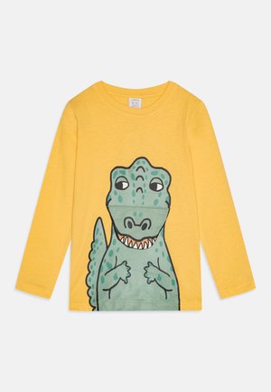 T-shirt jaune à manches longues présentant un motif de dinosaure vert avec des contours noirs, une expression souriante et des détails texturés sur le corps.