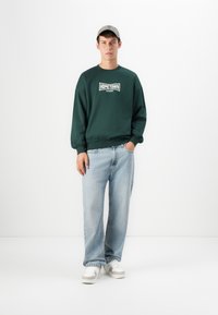 Young man wearing green sweatshirt with "Hometown Champion" text, light blue jeans, white sneakers, and a beige cap, standing against white background.