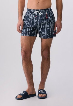 Uomo che indossa pantaloncini da bagno blu navy con motivo a corda bianca e coulisse bianca, abbinati a sandali slip-on blu navy con logo bianco.