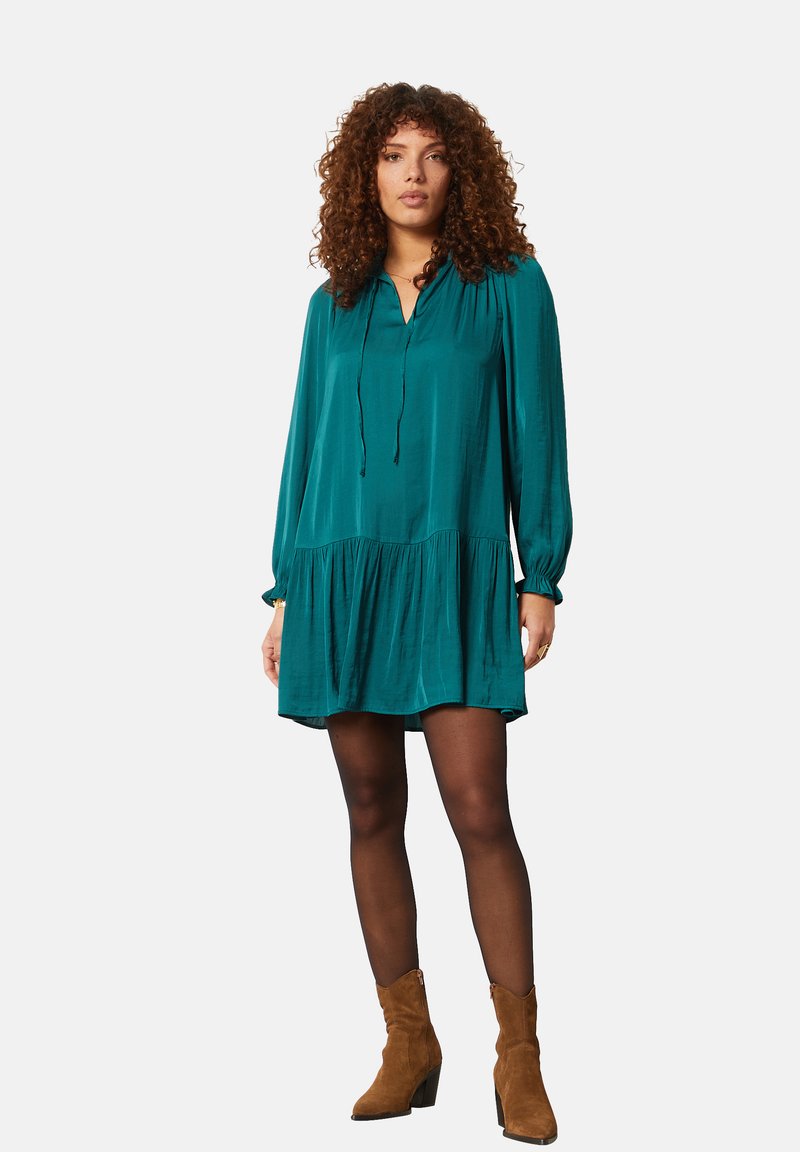 Teal, knielange jurk met een trekkoordhalslijn, lange mouwen en een uitlopende onderkant. Gecombineerd met tan enkellaarsjes.