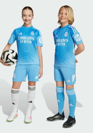 Kaksi lasta hymyilee, pukeutuneina vaaleansinisiin Real Madridin jalkapallopaitoihin, toinen pitää mustavalkoista jalkapalloa kädessään, seisten yksinkertaista taustaa vasten.