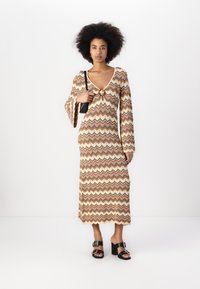 Robe en tricot beige avec des motifs en zigzag orange, marron et bleu, dotée d'un décolleté en V profond, de manches évasées et d'un détail central accentué.