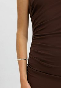 Bar arm iförd ett silverarmband bredvid en rynkad mörkbrun ärmlös klänning.