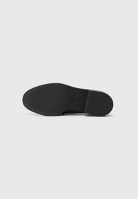 Semelle de chaussure noire avec une texture lisse, dotée de rainures horizontales pour une meilleure adhérence et d'une pointe arrondie. Design minimaliste avec un logo discret.