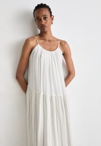 Weißes plissiertes Kleid mit dünnen Trägern, rundem Ausschnitt und schwarzen vertikalen Streifen. Fließender Stoff mit einem gestuften Saum. Weiche Textur.