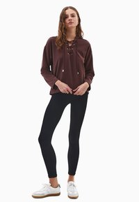 Brązowa bluzka z długimi rękawami i sznurowanym dekoltem, zestawiona z czarnymi legginsami i białymi sneakerami, cechująca się luźnym krojem i gładką teksturą materiału.