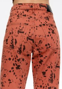 Oranžinės džinsai su juodu purvo raštu, su aukštu liemeniu, penkiomis kišenėmis ir juodu logotipu nugaroje juosmens srityje.