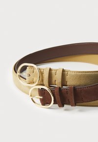 Deux ceintures en daim beige et en daim marron, chacune avec des boucles circulaires en ton or, montrant des textures et des détails de couture distincts.