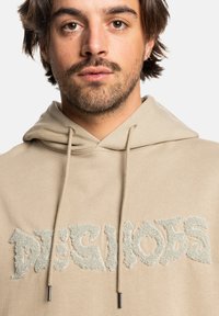 Beige hoodie with a textured, light gray logo reading "DESHOES." Features a kangaroo pocket and adjustable drawstring hood. Soft fabric texture.