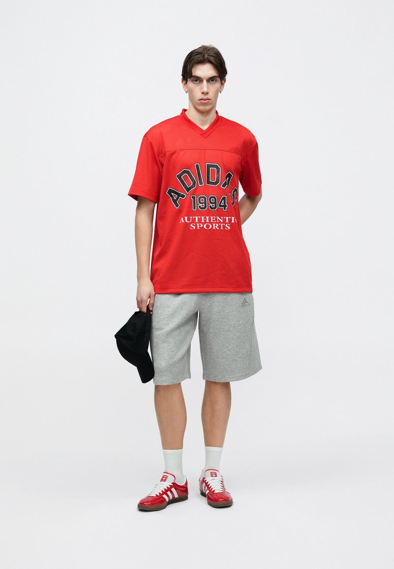 Jeune homme debout portant un maillot de sport Adidas rouge, un short Adidas gris, des chaussettes blanches, des baskets Adidas rouges, et tenant une casquette noire dans une main.